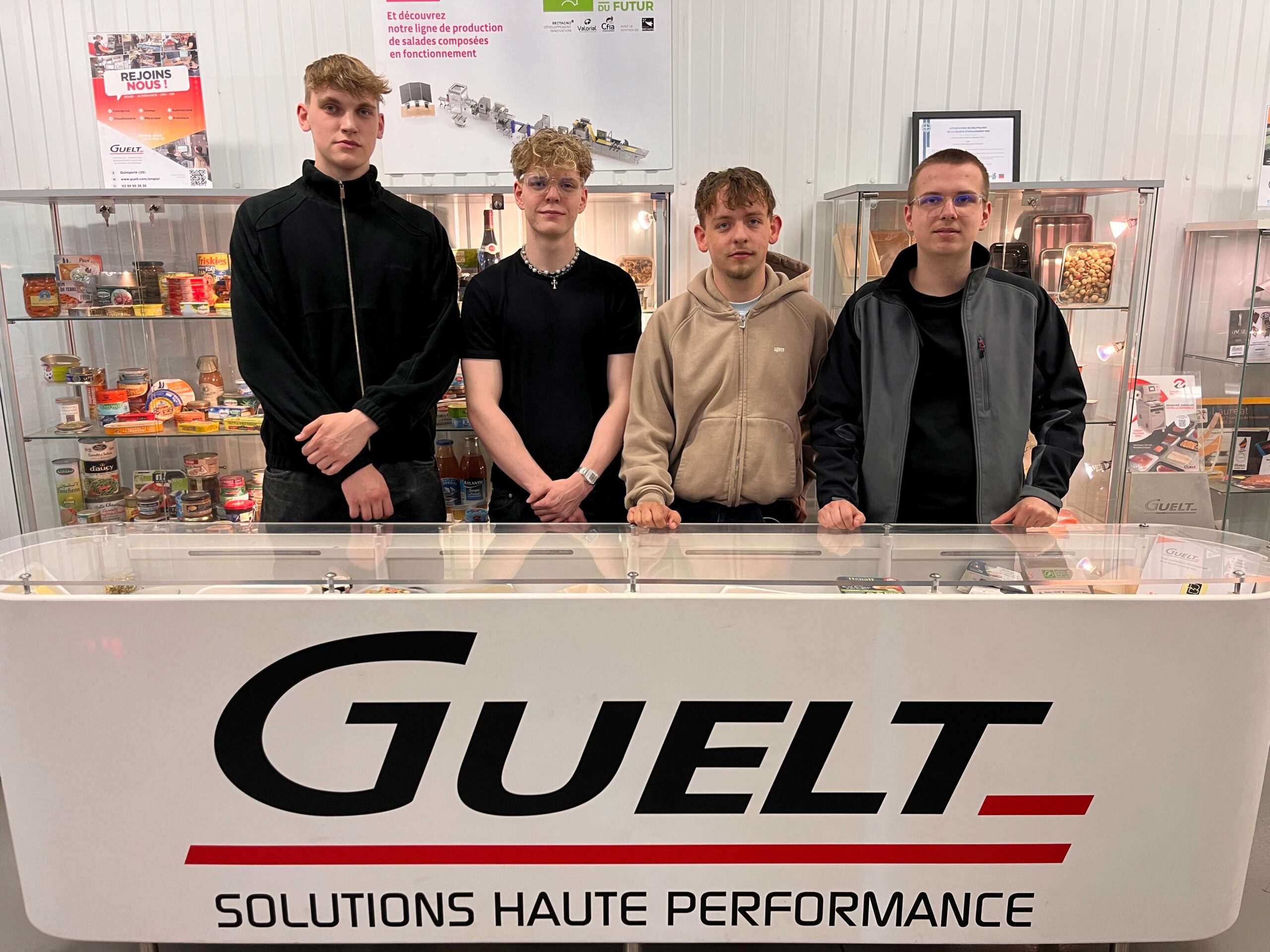 Die vier deutschen Austausschüler bei der Firma Guelt. Sie stehen hinter einem Tresen mit der Aufschrift Guelt und blicken in die Kamera.