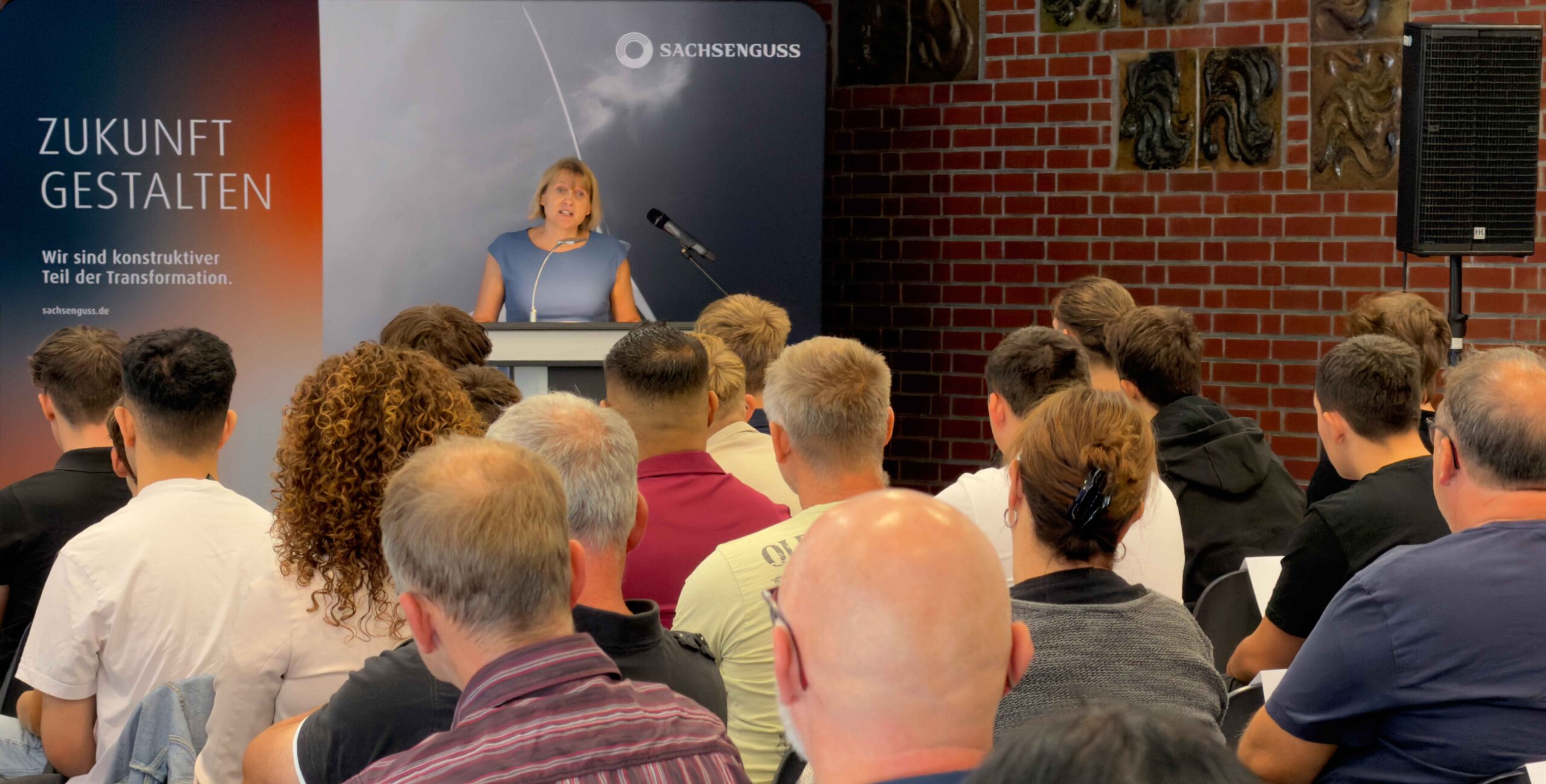 Die Personalleiterin von Sachsen Guss steht an einem Rednerpult vor einer Gruppe von Zuhörern. Sie begrüßt die neuen Auszubildenden und ihre Eltern.