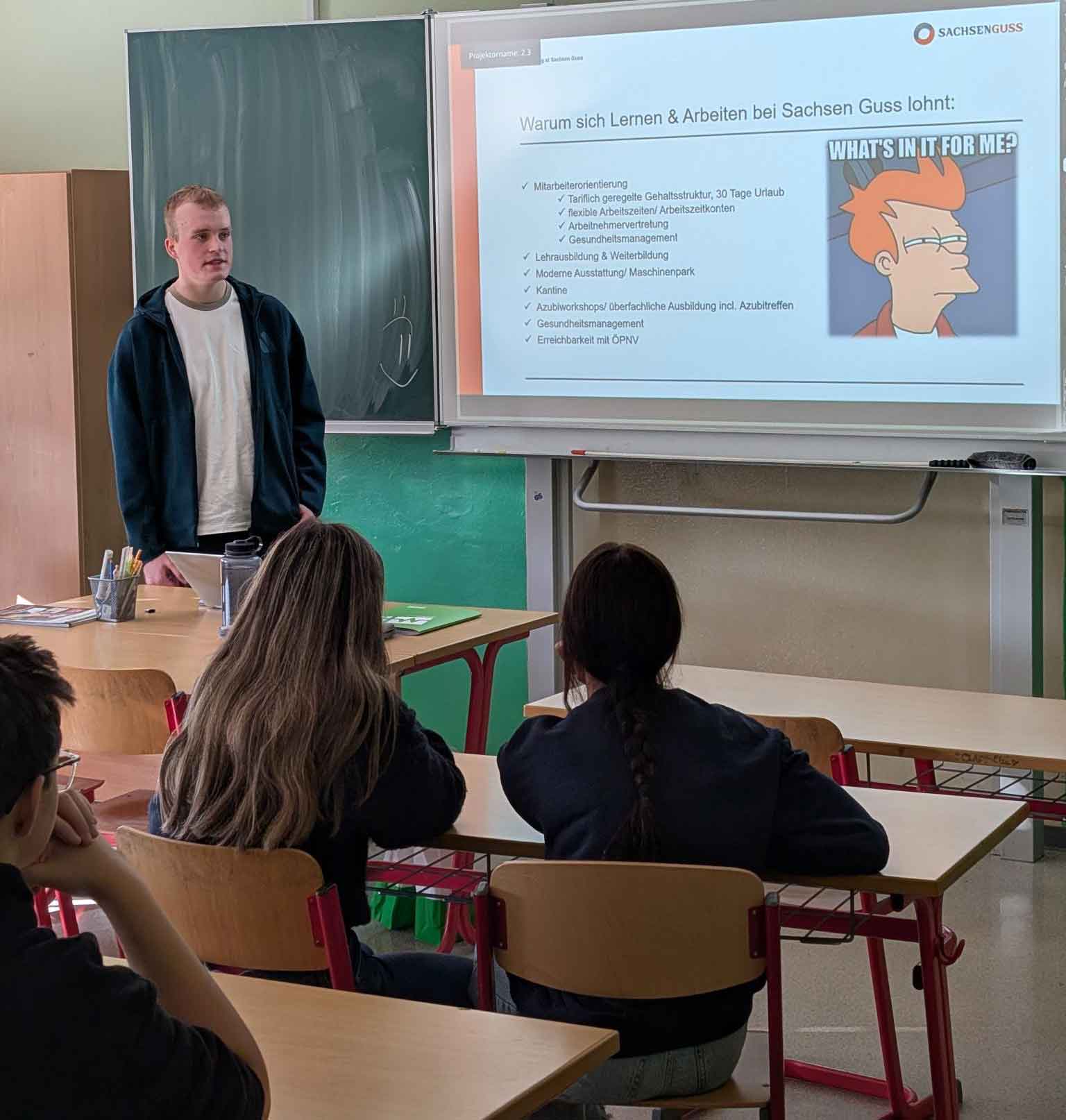 Lukas Hofmann steht in einem Klassenzimmer vor einer Schuleklasse. Neben ihm ist ein Slide einer Power-Point-Präsentation geöffnet. Überschrift auf dem Slide: Warum sich Lernen & Arbeiten bei Sachsen Guss lohnt.