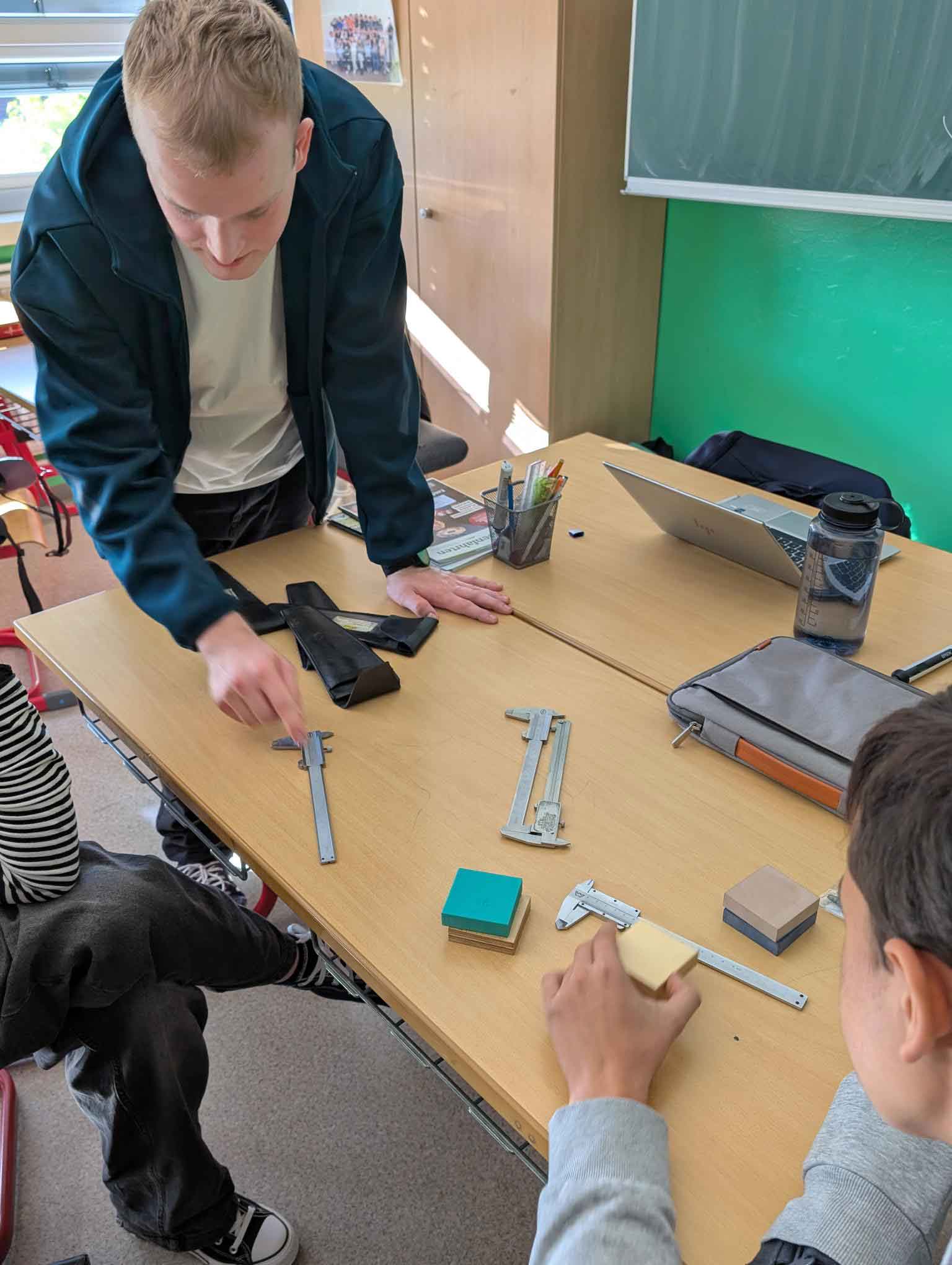 Lukas Hofmann steht in einem Klassenzimmer an einem Tisch, an dem ein Schüler und eine Schülerin sitzen. Er deutet auf einen Messschieber, der auf dem Tisch liegt.